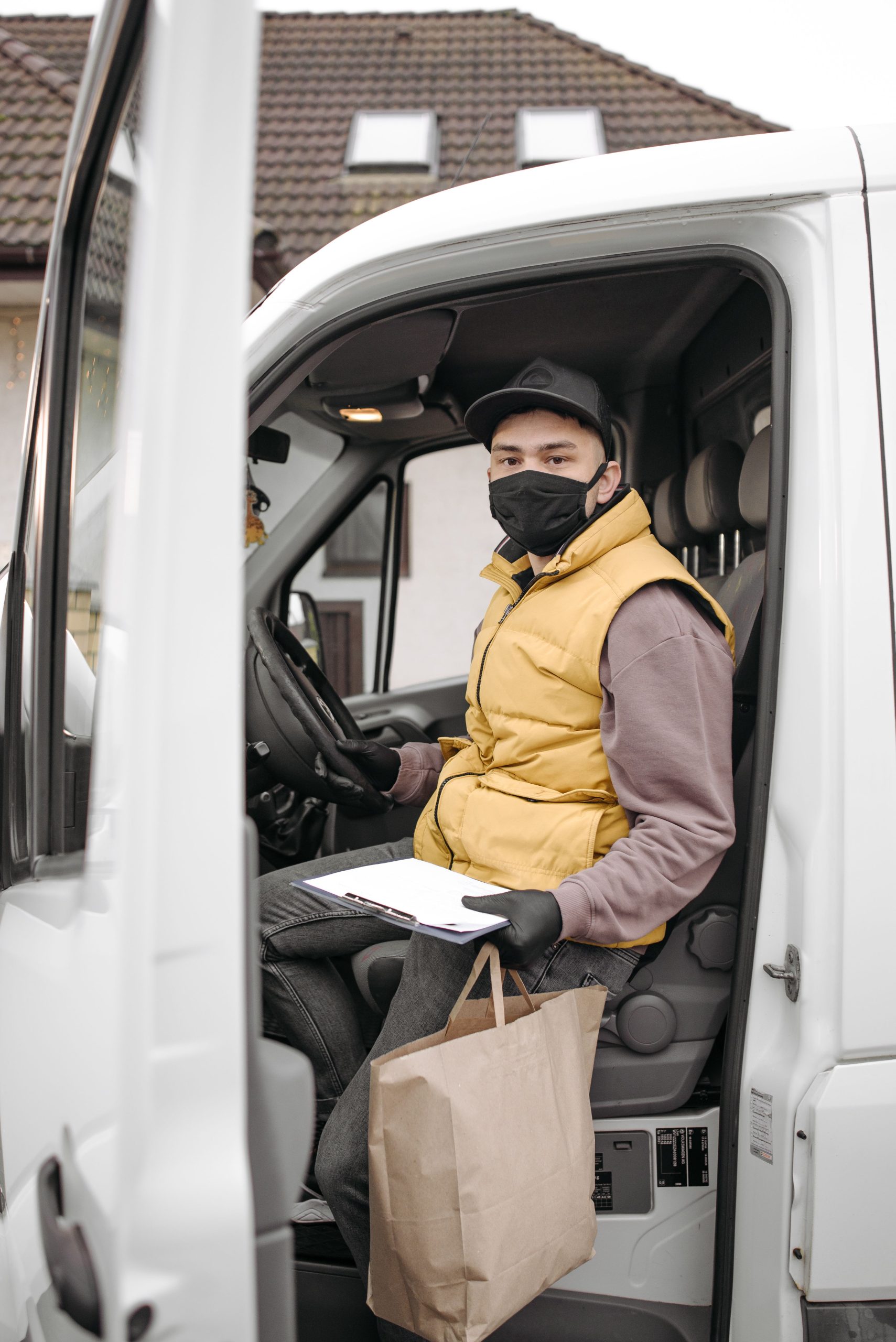 A delivery driver wearing a face mask sits inside a van holding a paper bag and clipboard, symbolizing contactless delivery